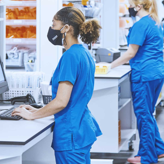 empower-pharmacy-technician-operating-a-computer-system-550x550.png Woman in blue scrubs and a black mask standing and working at a computer.