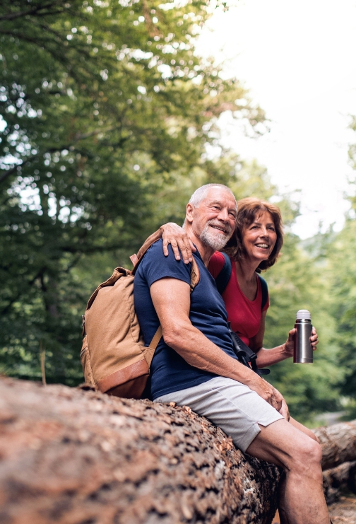 An older aged man and woman sitting arm and arm outdoors on a fallen tree.