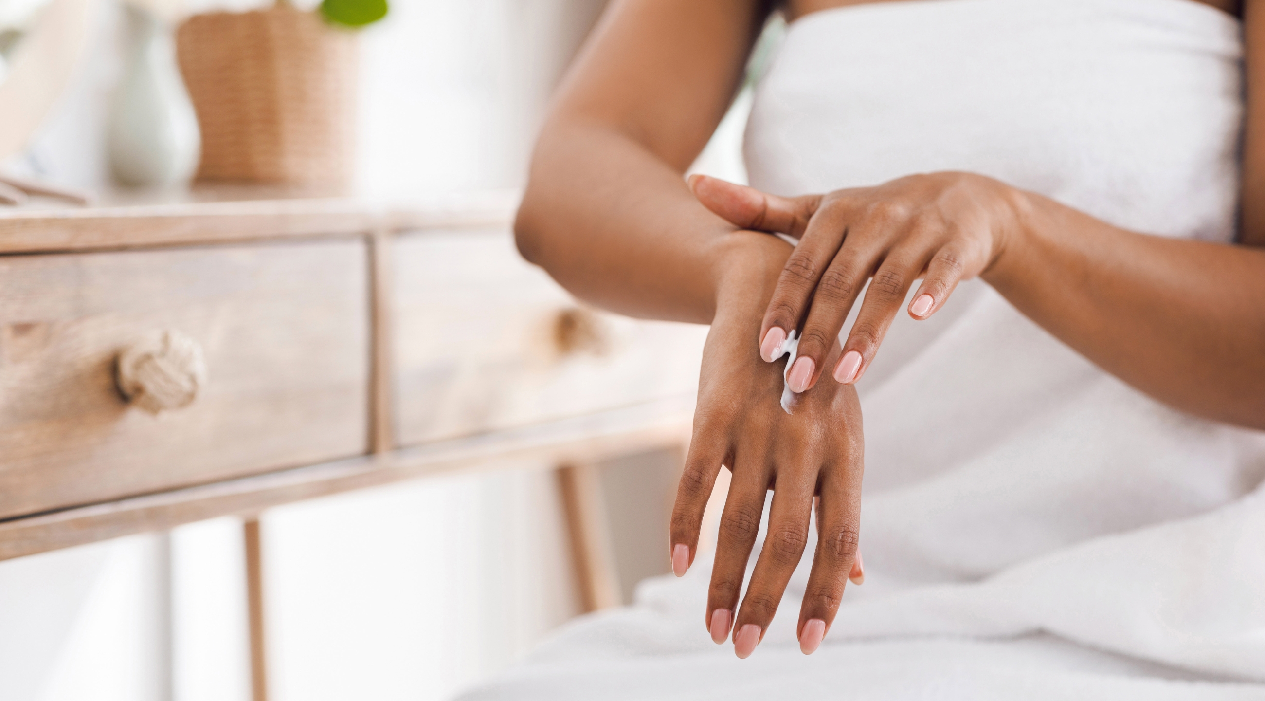 An African American woman applying lotion to her hands in a white bath towel seated at a desk.