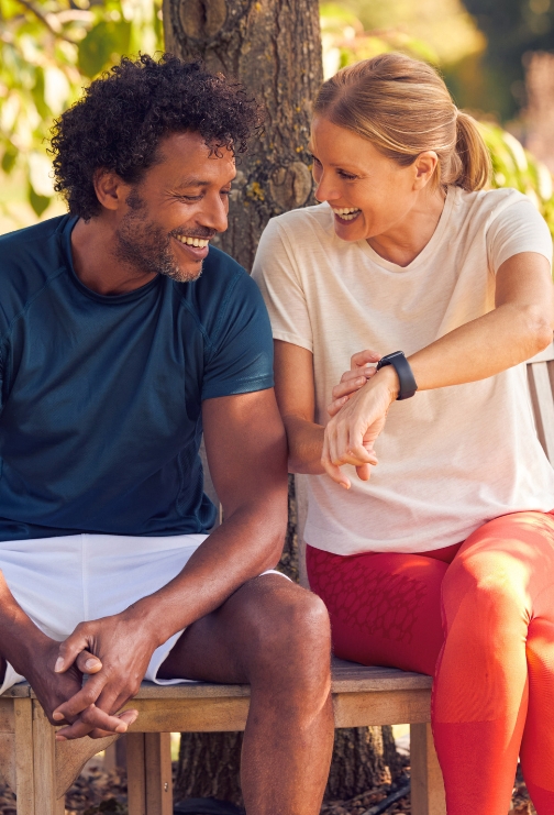 A woman with blonde hair showing her smart watch to an African American man while both sitting on a bench outside.